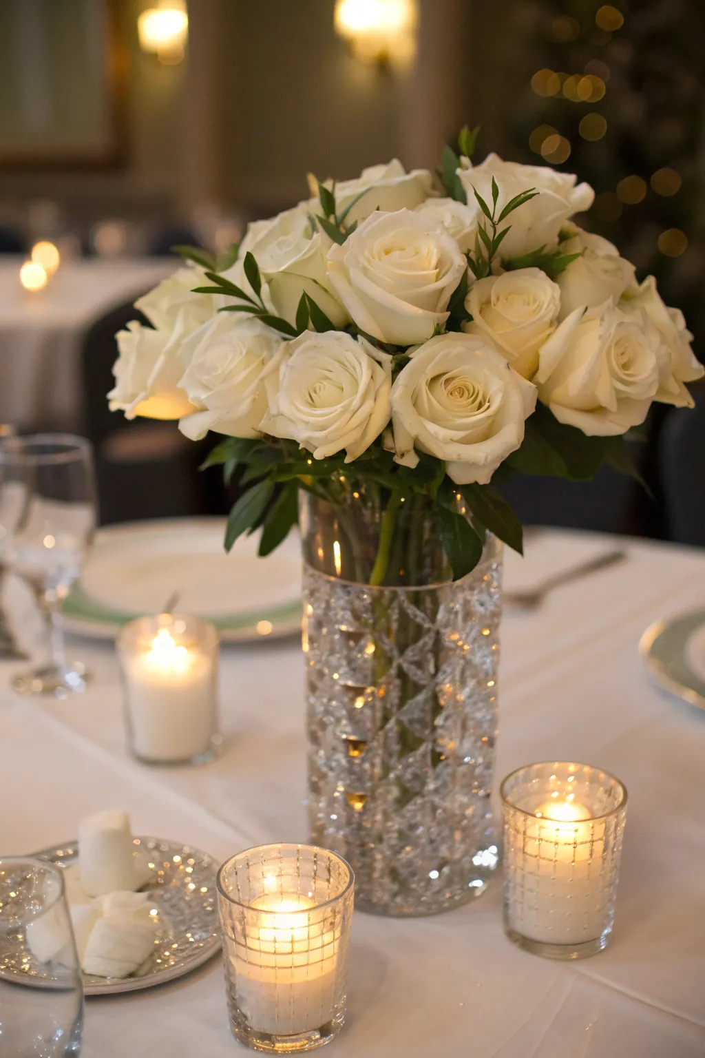 Elegant centerpiece with white roses in a crystal vase surrounded by flickering votive candles.