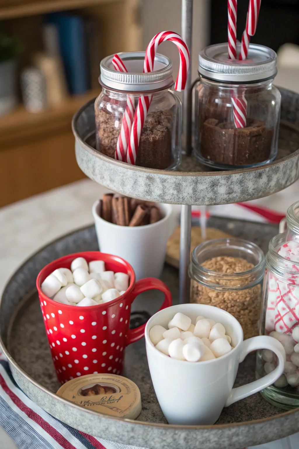 Tiered tray styled with mugs, cocoa jars, candy canes, and marshmallows for a festive cocoa bar.