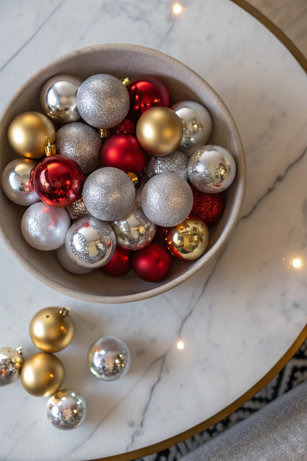 A shallow bowl filled with shiny silver, gold, and red ornaments on a marble coffee table.