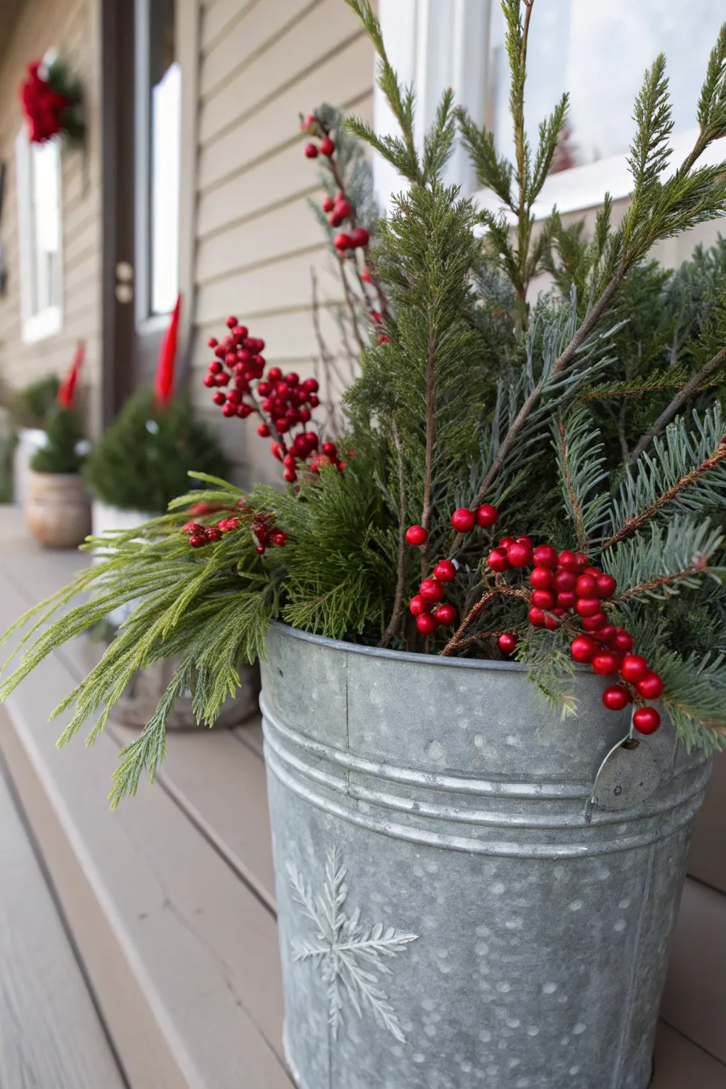 Galvanized metal bucket planter filled with pine, cedar sprigs, and red berries beside a wooden porch wall.