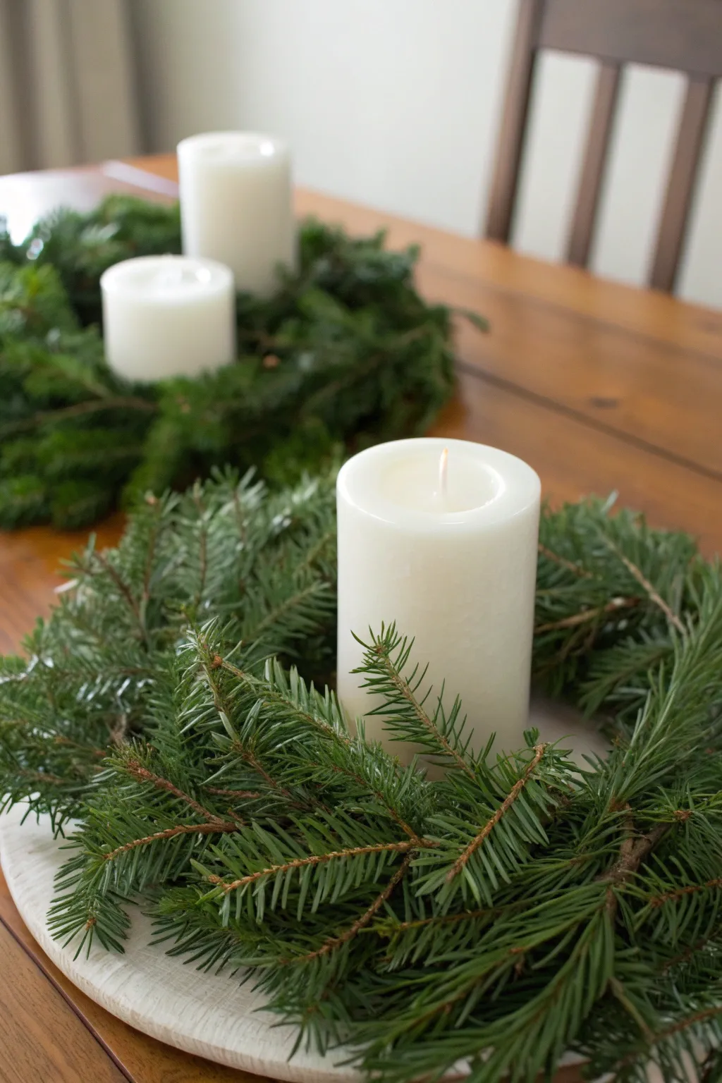 Evergreen garland ring surrounding white pillar candles on a wooden table.