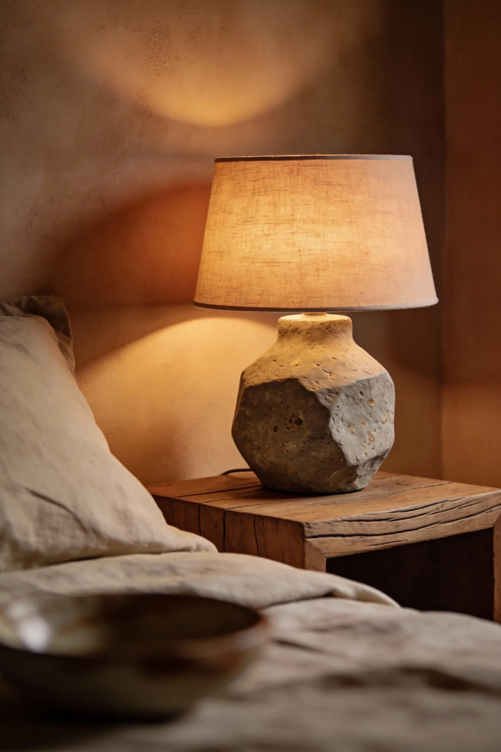 Stone or clay table lamp with fabric shade on a wooden nightstand in a warm bedroom.