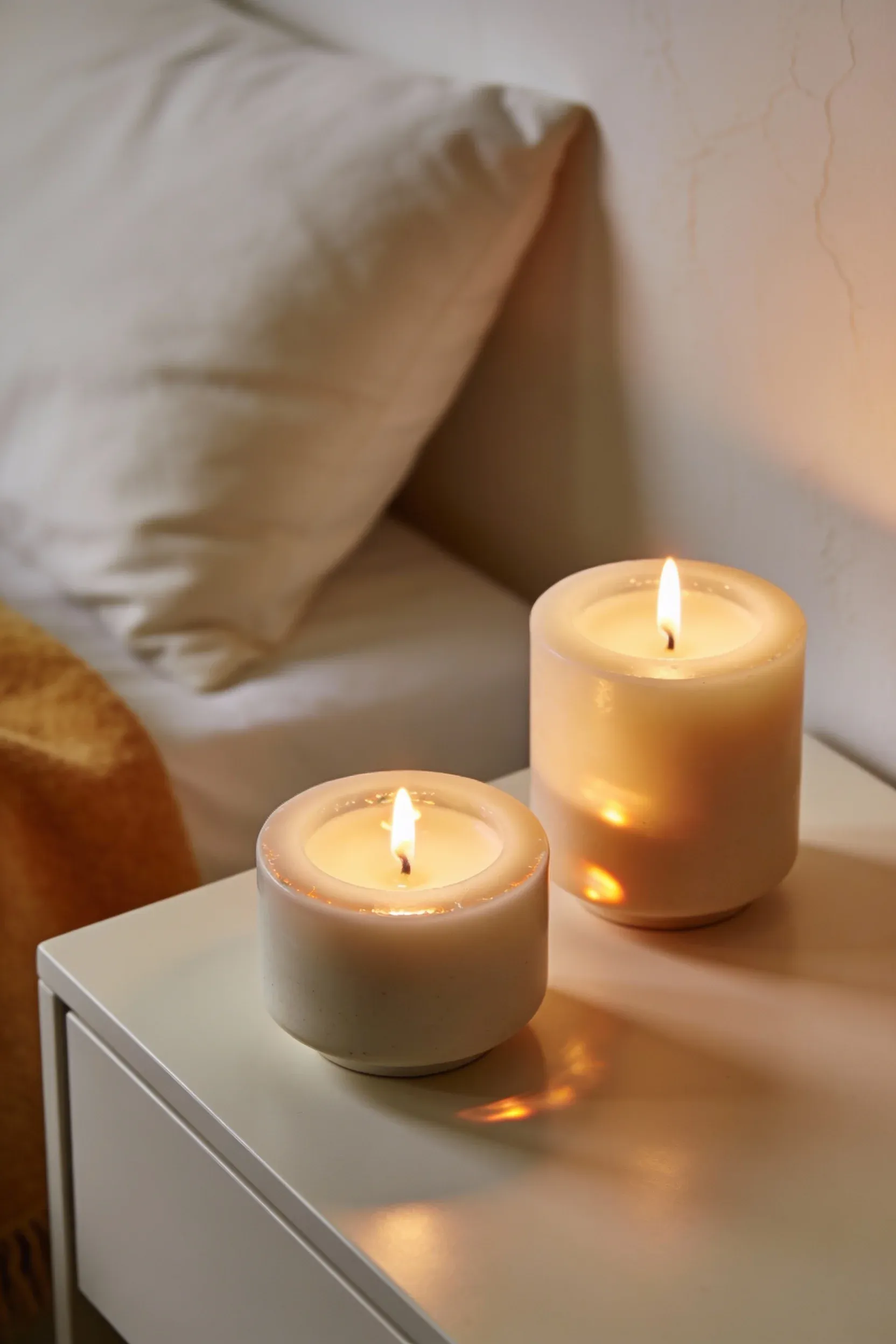 Cream and beige scented candles glowing softly on a nightstand in a white and beige bedroom.