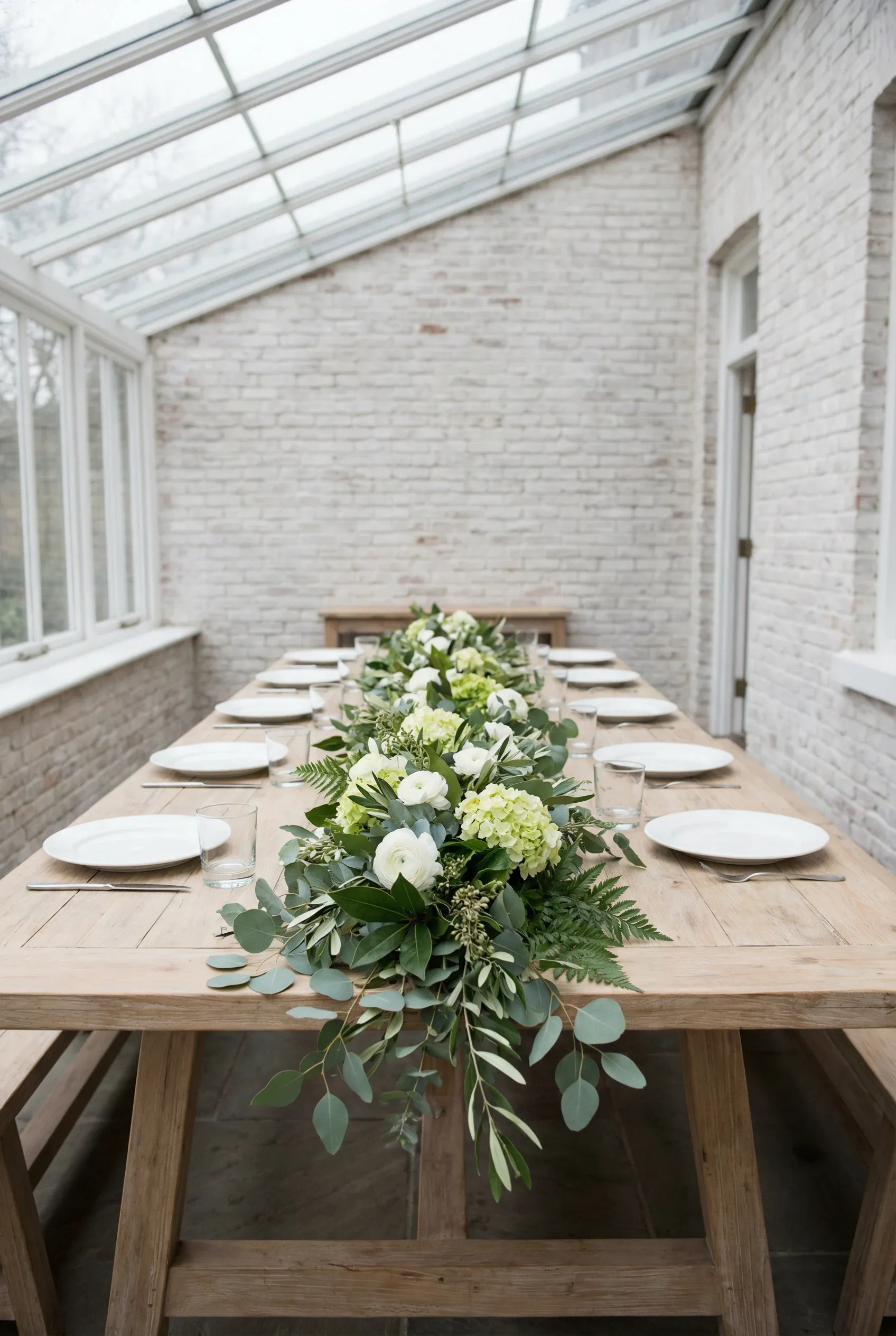 A low greenery and white flower runner extending down the center of a light wood dining table.
