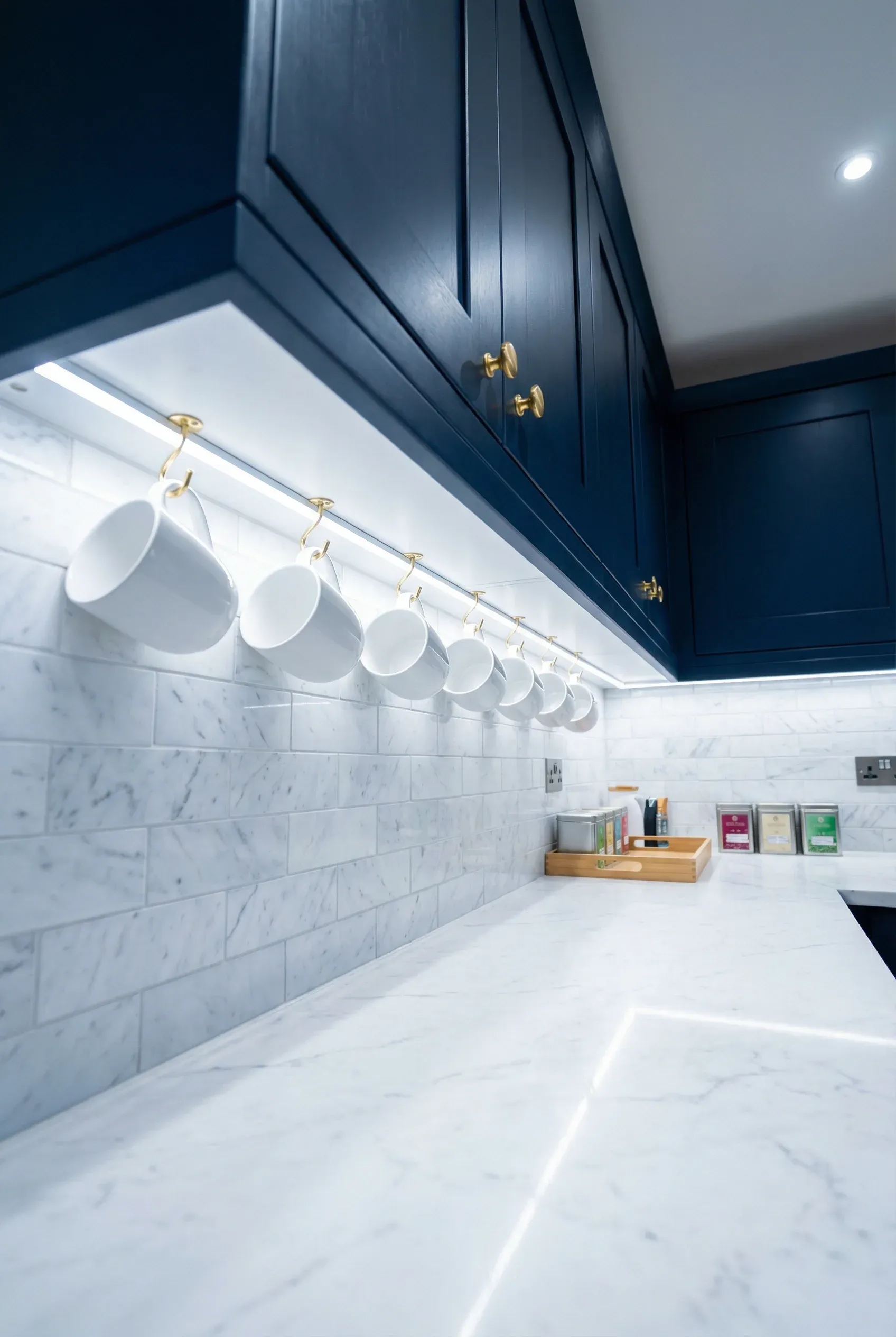 Brass hooks under navy blue kitchen cabinets holding a row of white ceramic mugs over a marble counter.