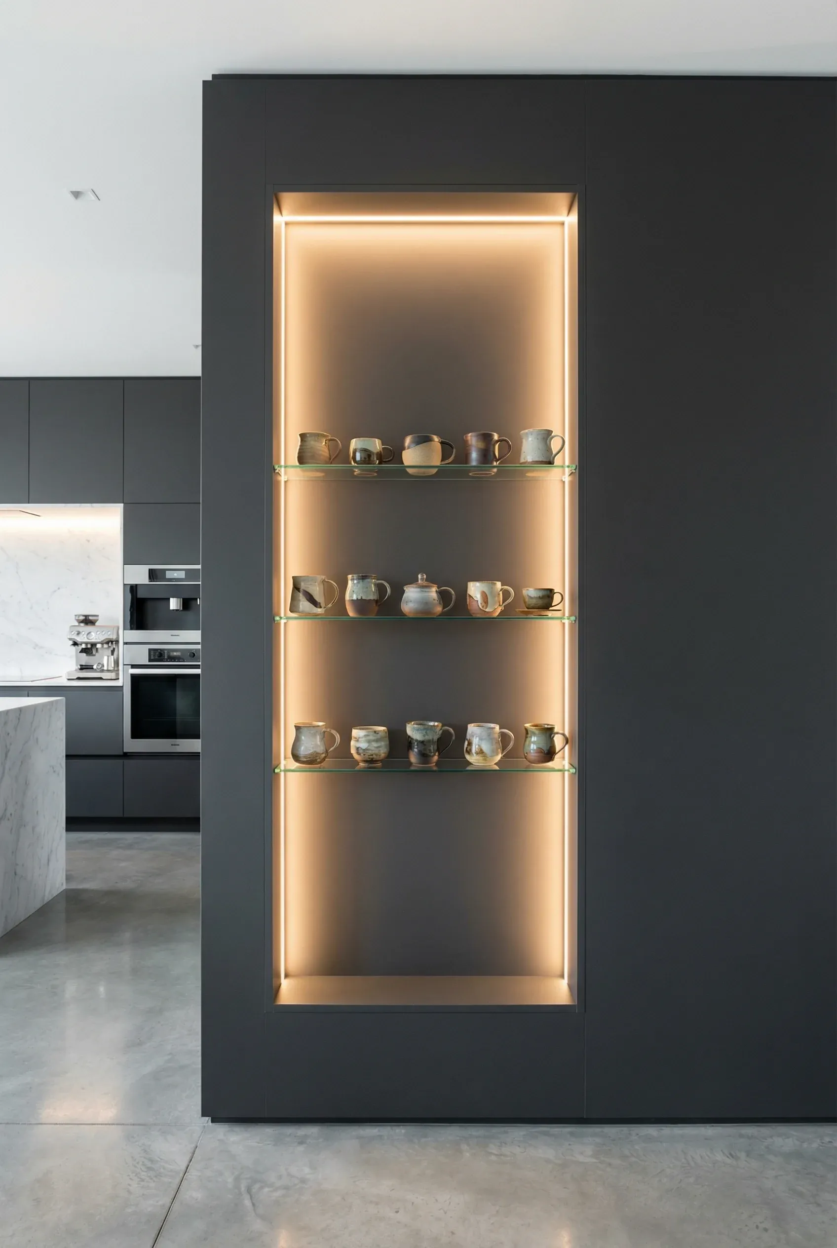 A modern kitchen wall with a glowing recessed niche displaying artisanal ceramic mugs on glass shelves.