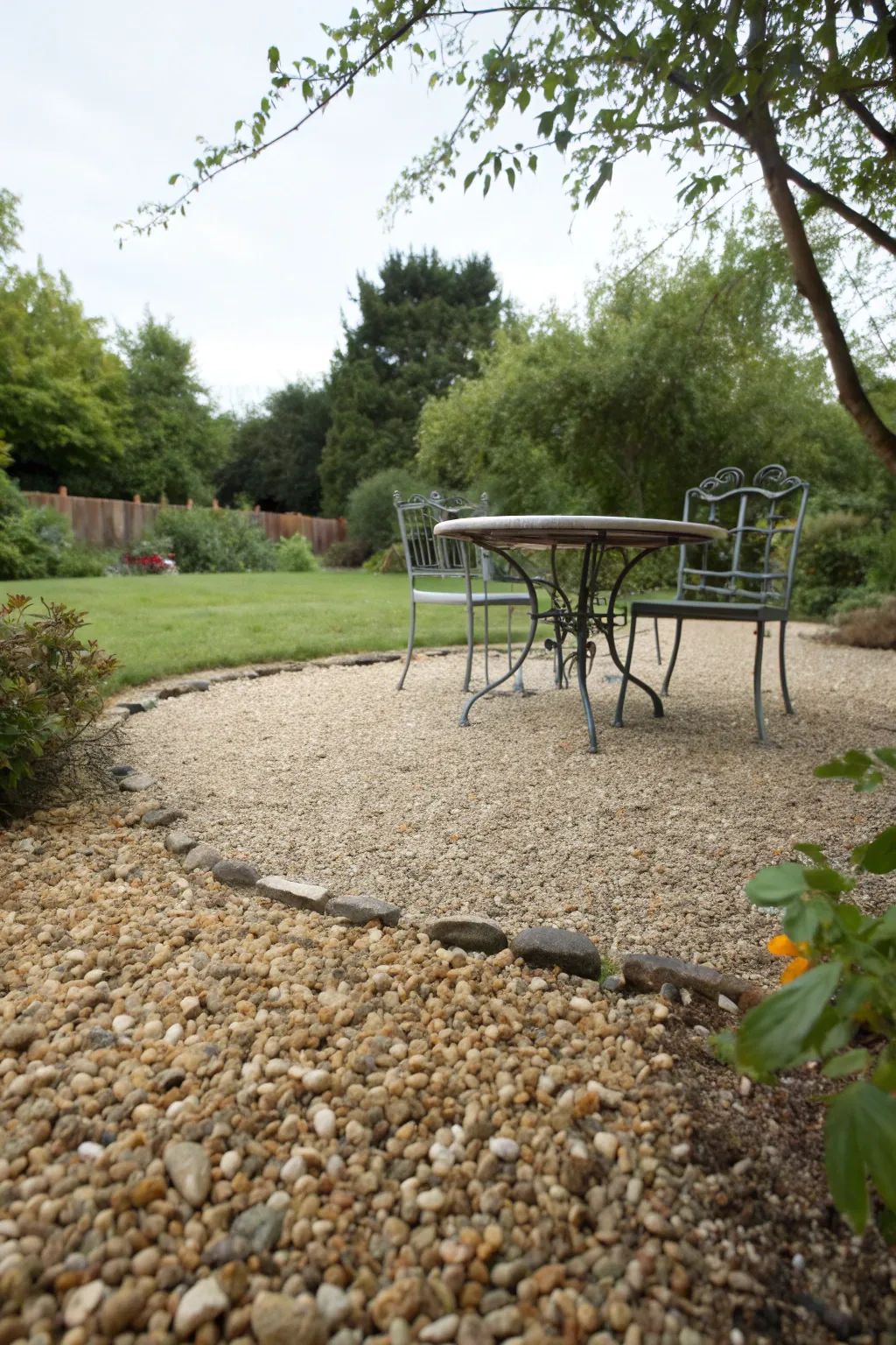 A black wrought iron dining set stands on a light tan pea gravel patio surrounded by hedges.