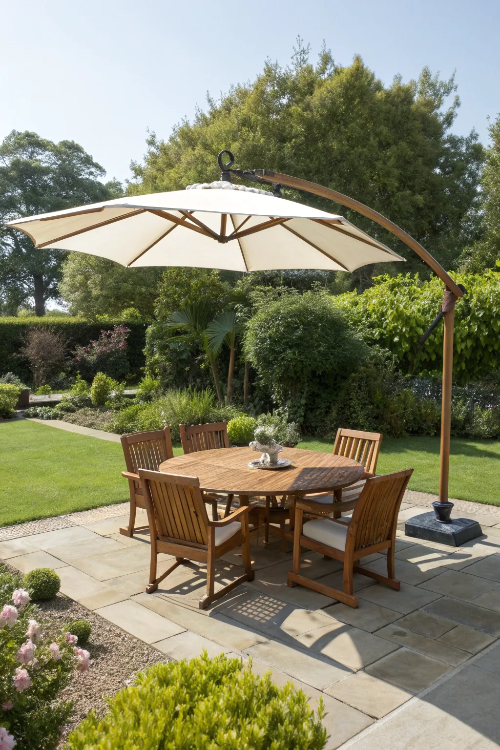 A large round teak table is shaded by a charcoal cantilever umbrella on a stone patio.