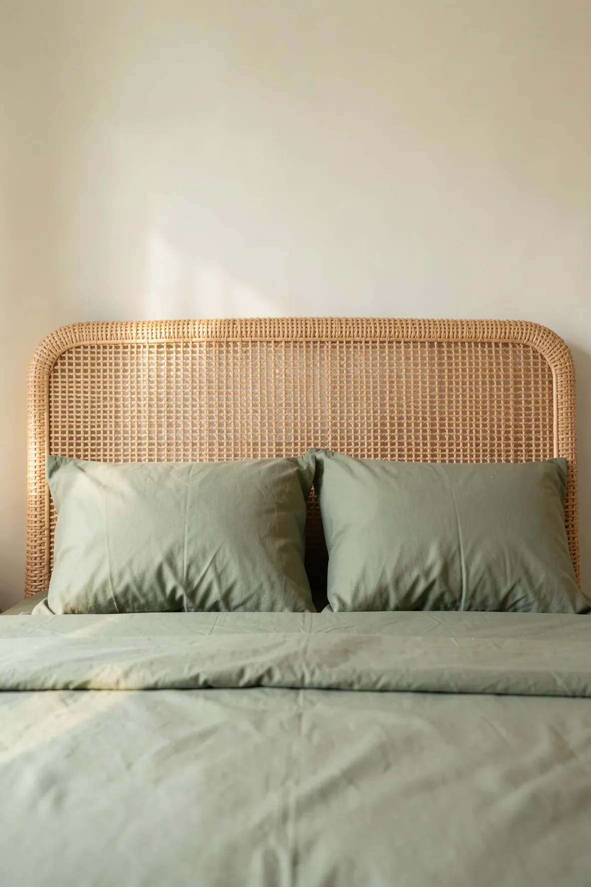 A woven rattan headboard with muted moss green sheets in a warm bedroom.