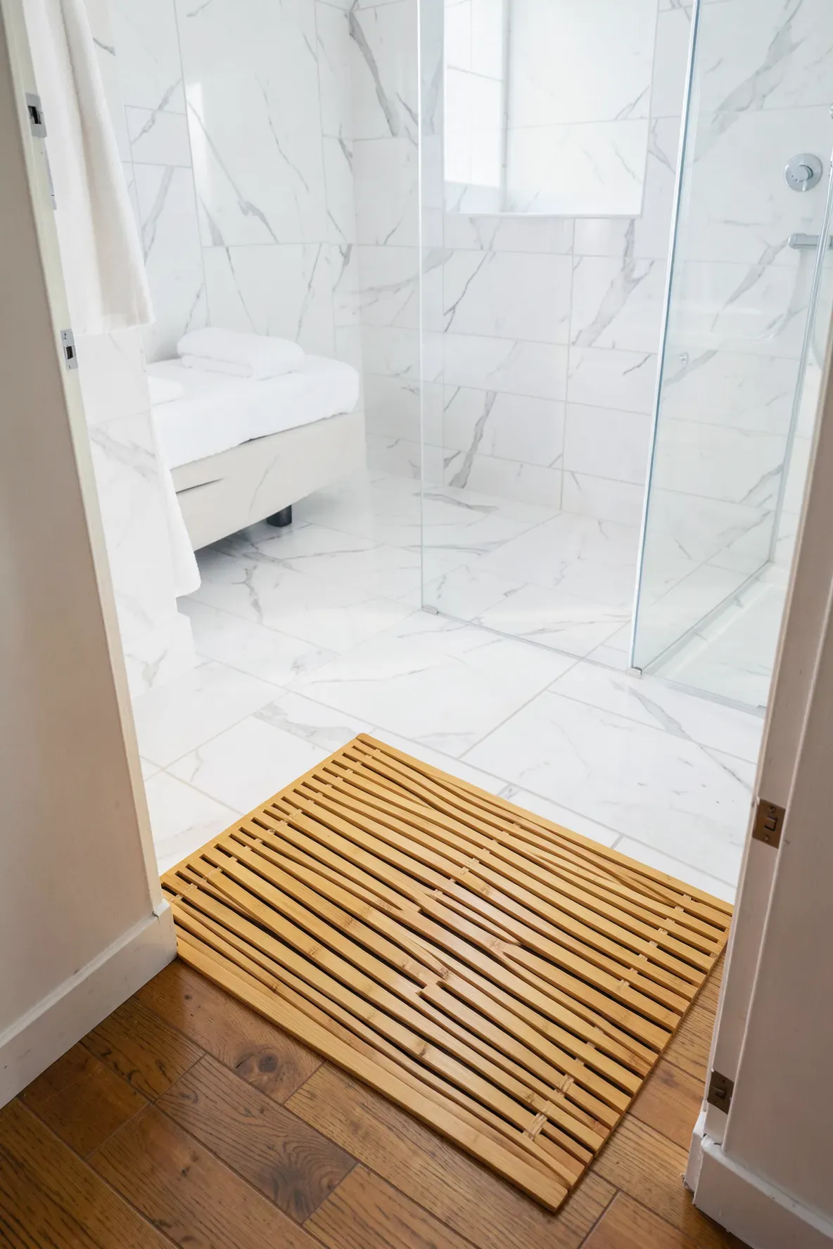 A bamboo slat mat on the floor between a bedroom and a bright marble en suite bathroom.