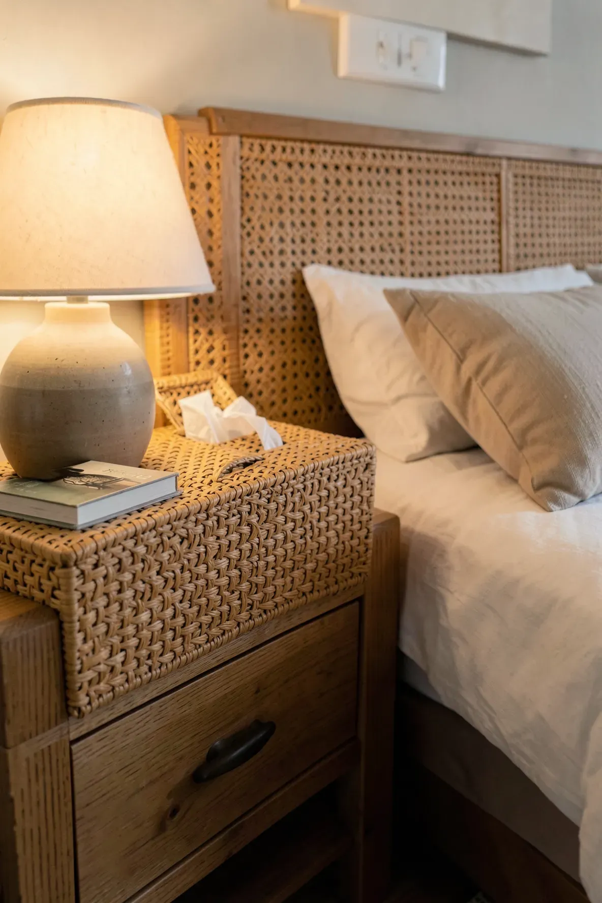 A decorative rattan tissue box cover on a wooden nightstand in a cozy, styled bedroom.