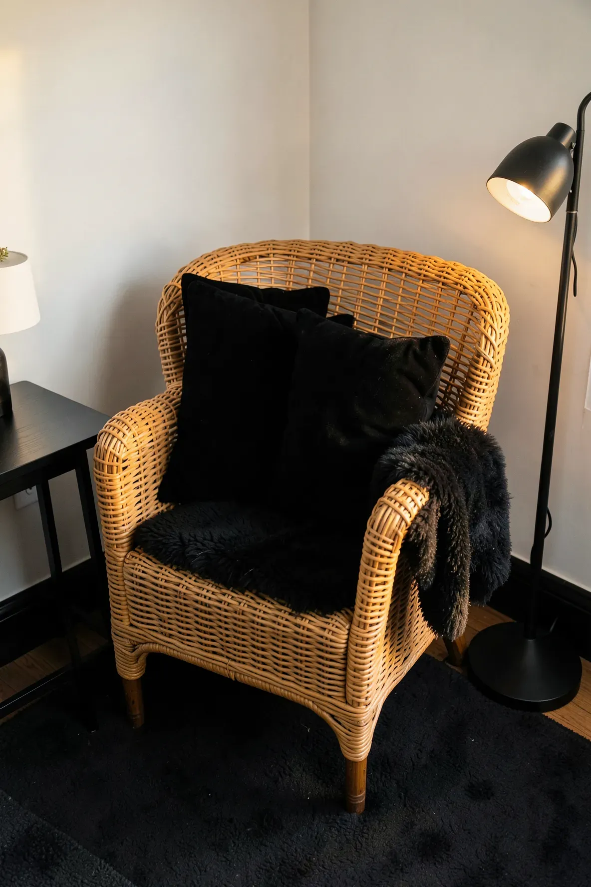 A woven rattan armchair in a bedroom corner with black velvet pillows and a black floor lamp.