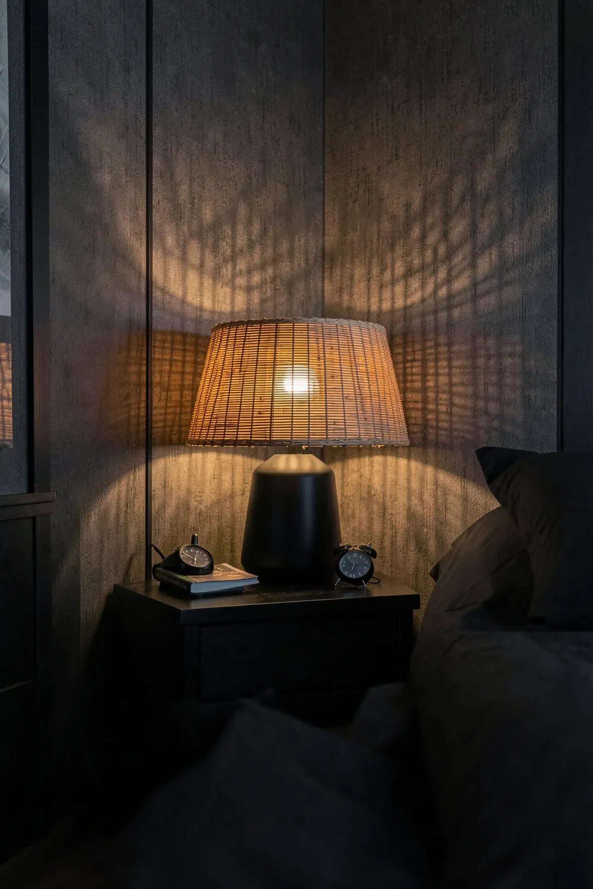 A black table lamp with a woven bamboo shade glowing warmly on a black nightstand.