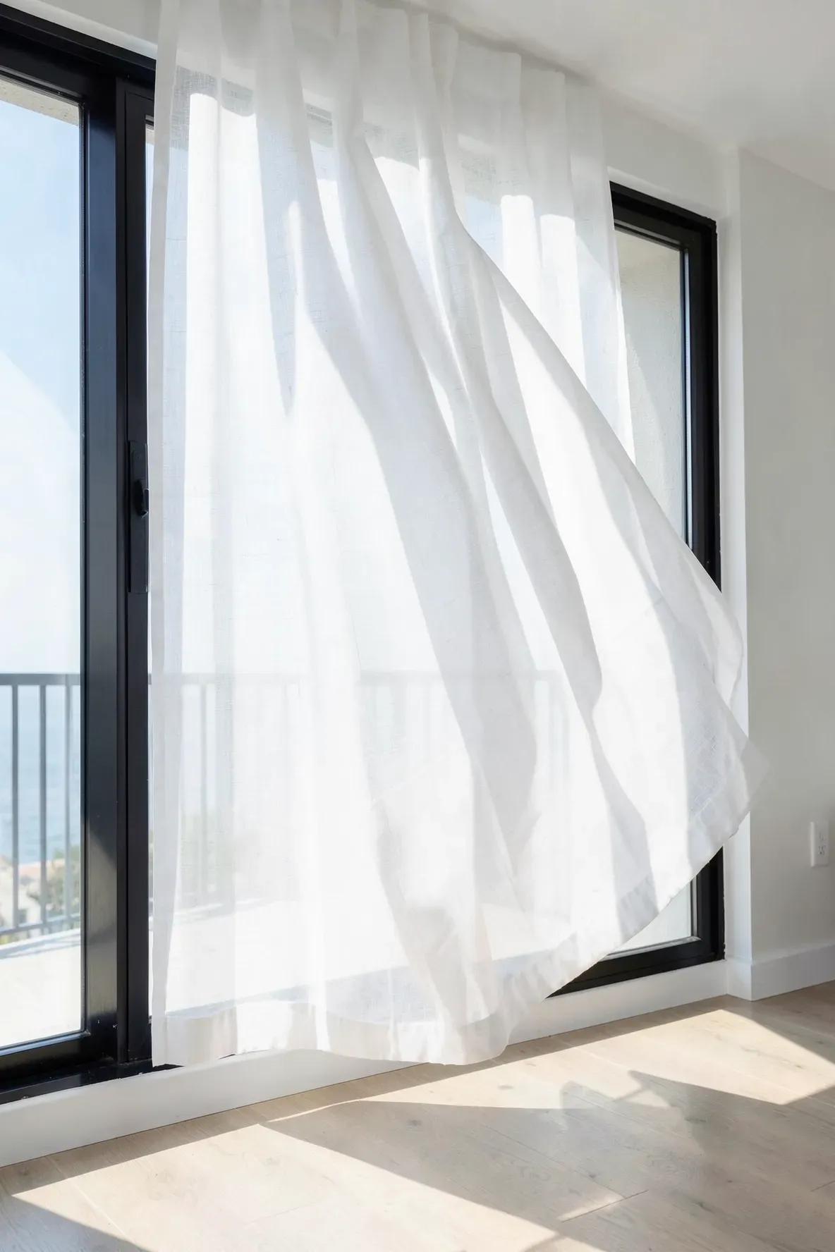 Thin, sheer white linen curtains fluttering in a breeze in front of a bright window in a beachy room.