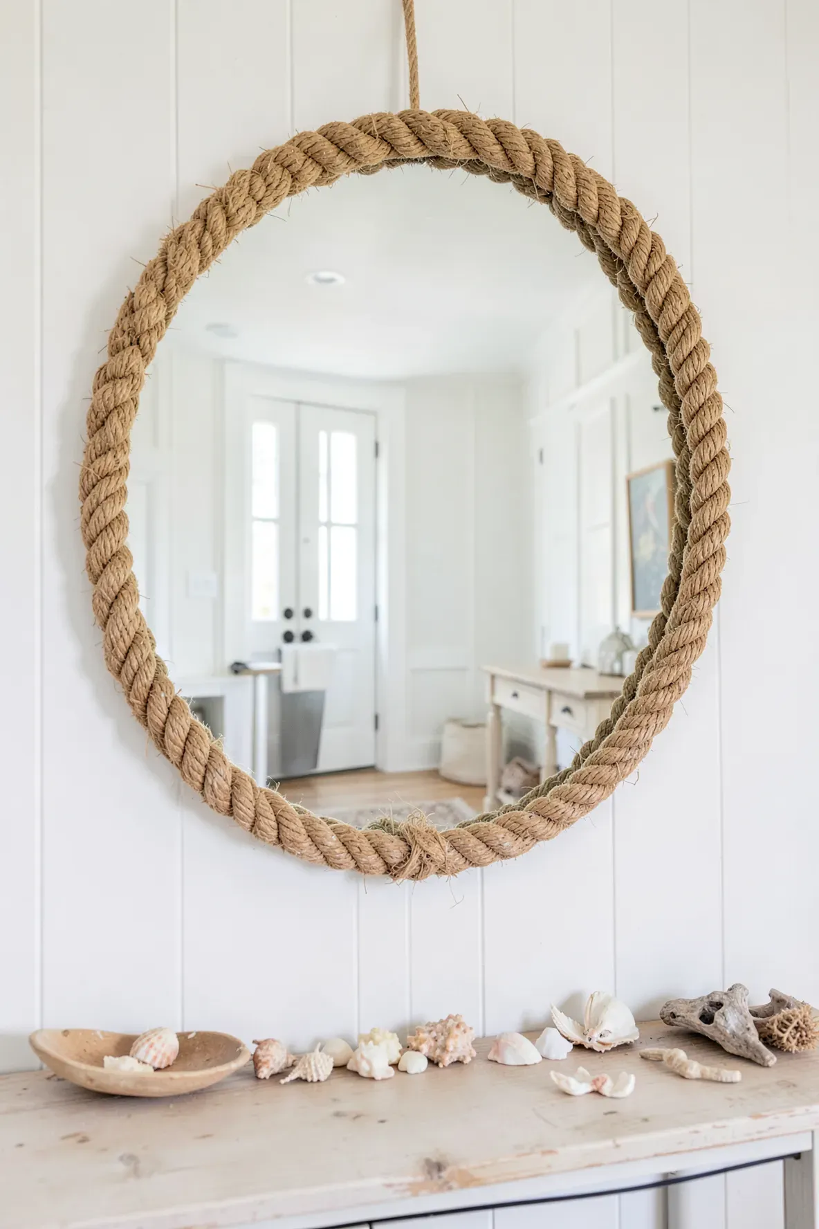 A round mirror with a thick rope frame hangs on a white wall, reflecting a bright beachy room.