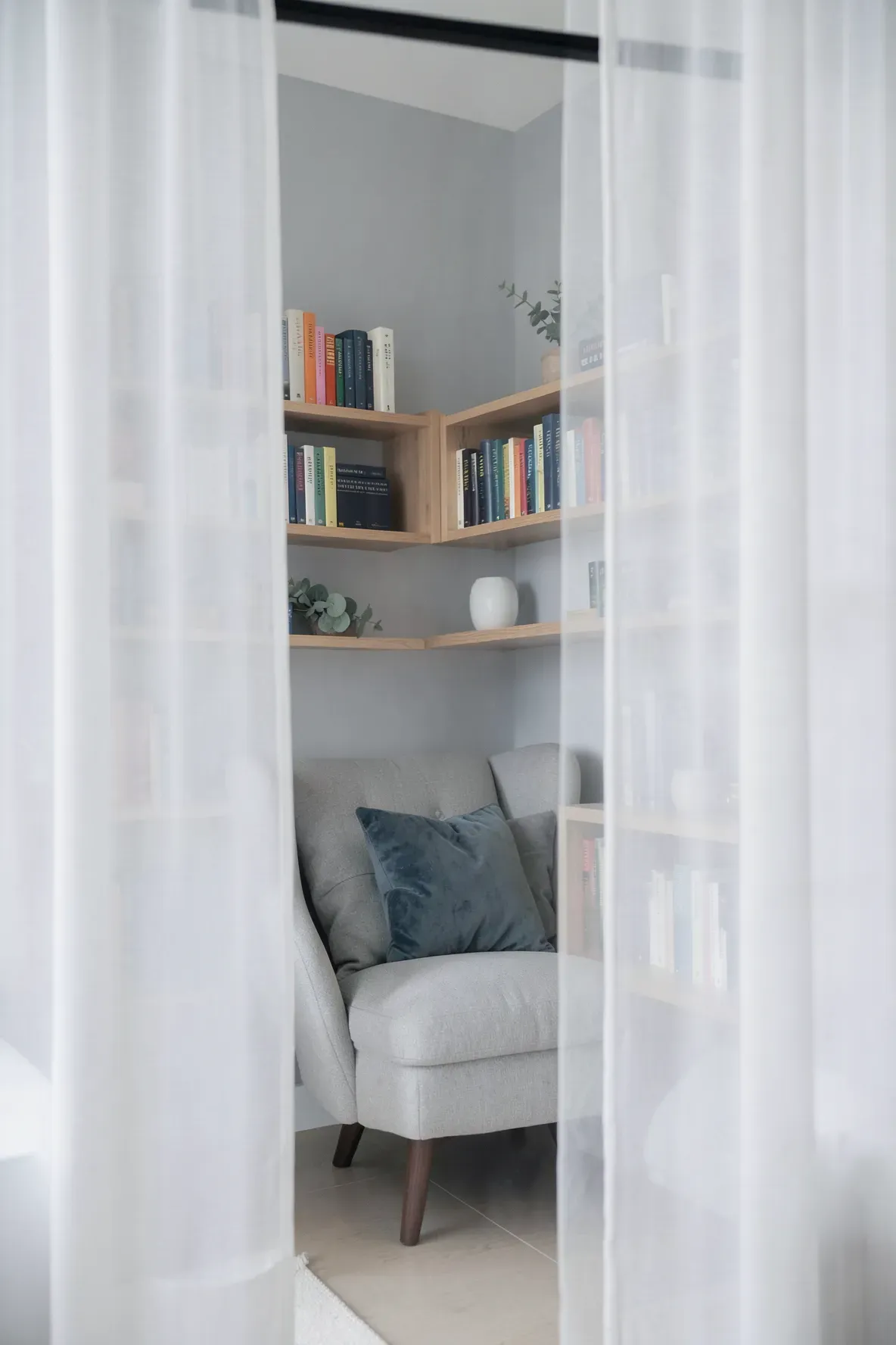 Sheer white curtains partially reveal a cozy reading chair and bookshelf in a quiet room corner.