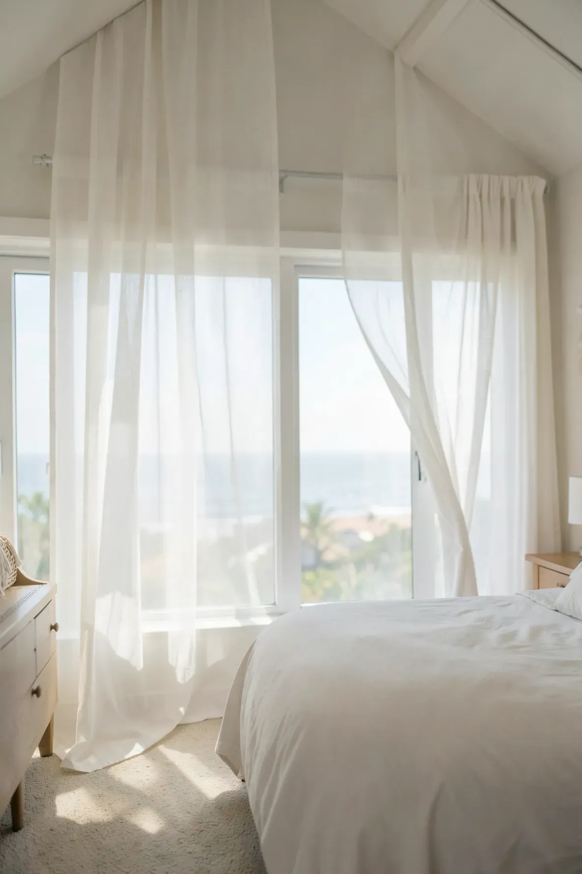 Lightweight white sheer gauze curtains filtering sunlight in a bright, airy coastal bedroom.