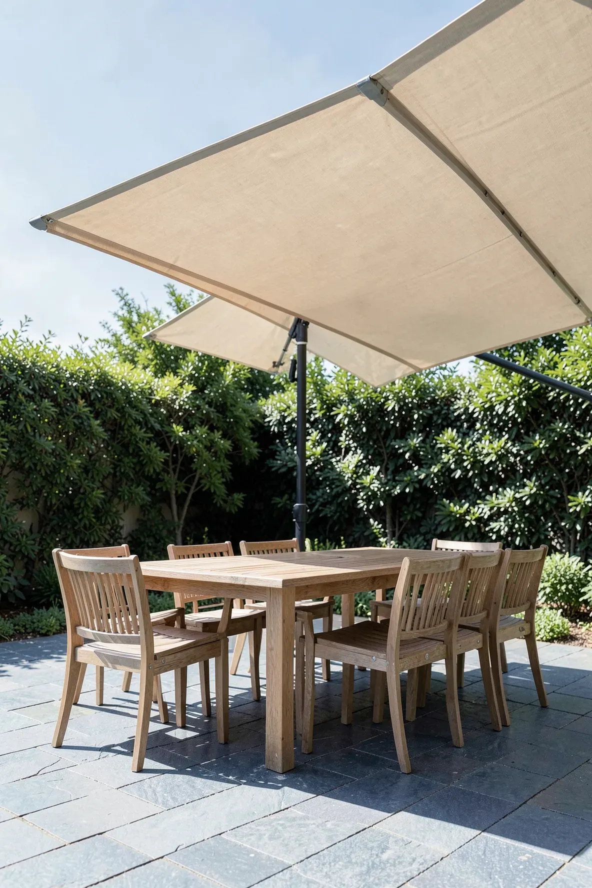 A large beige cantilever umbrella shading a teak dining table on a stone patio.