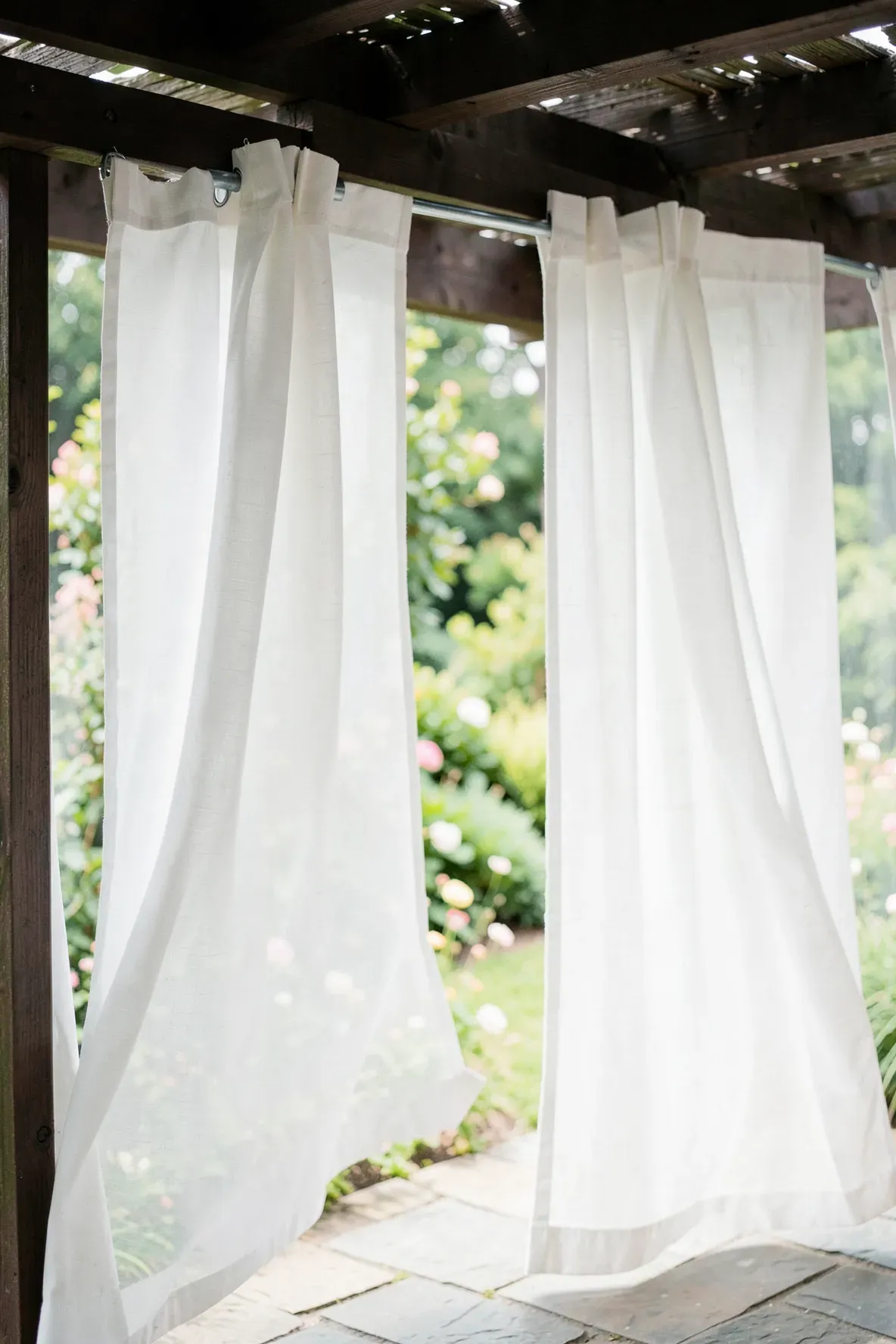 White weather-resistant curtains hanging from a wooden pergola on a private patio.