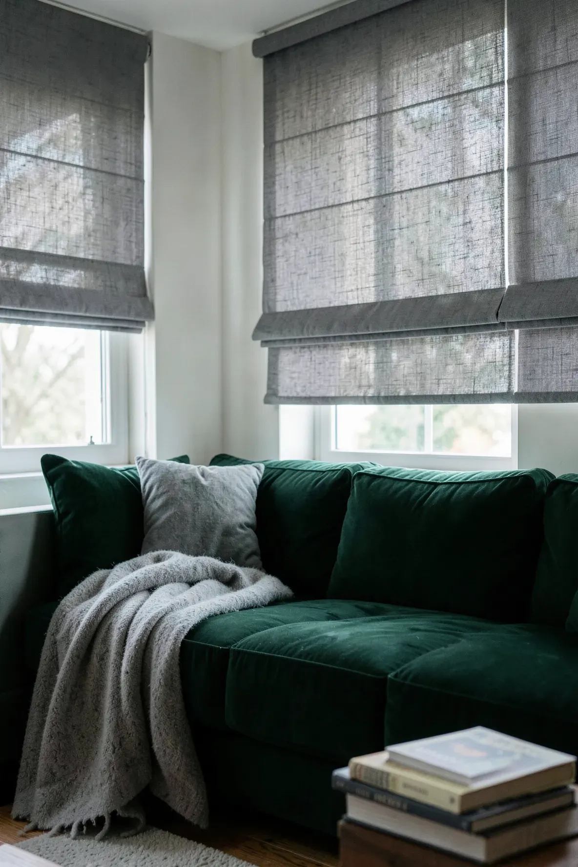 An emerald green sofa positioned in front of a window with textured grey linen Roman shades.