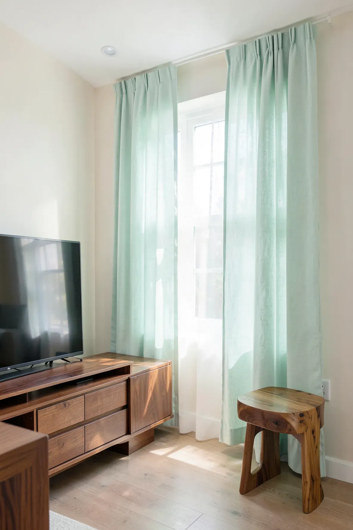 Soft mint curtains hanging next to warm walnut furniture in a bright, sunlit living room.