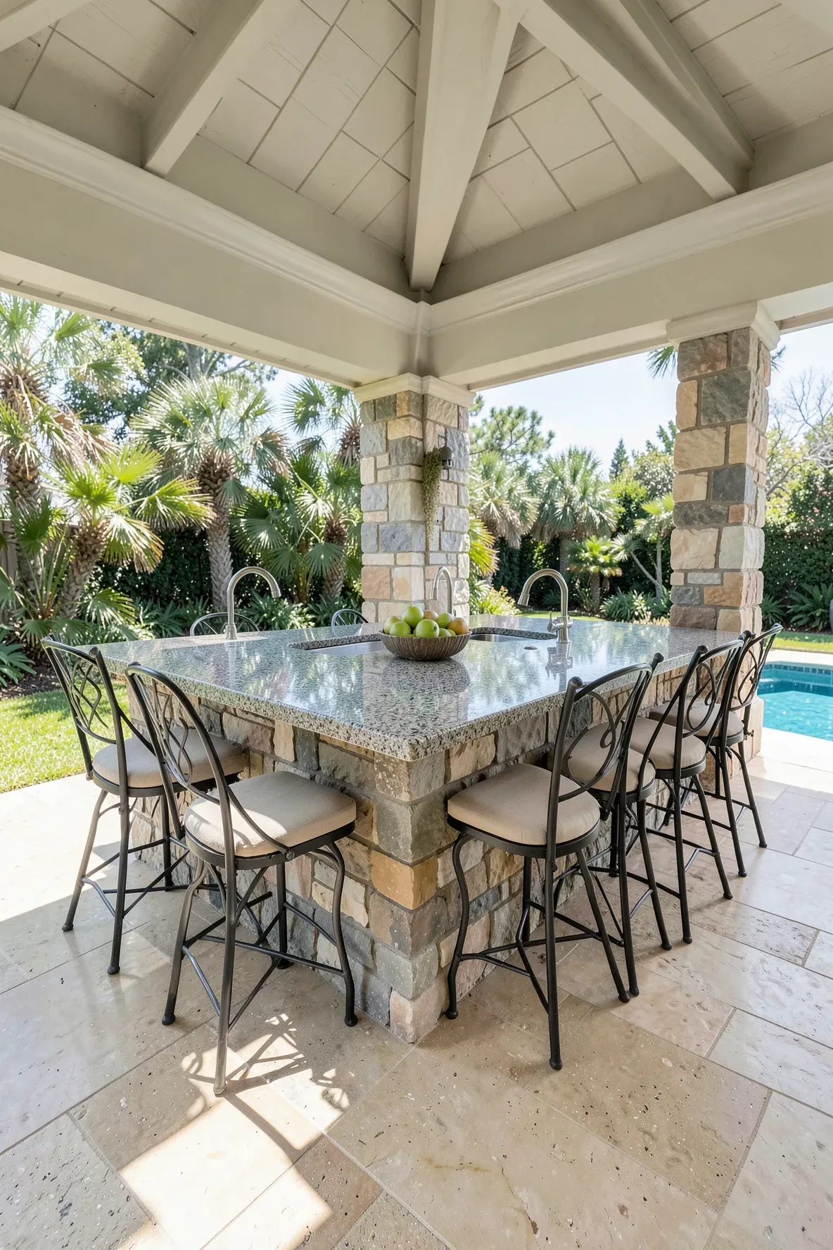 A large stone outdoor kitchen island with granite countertops and bar stools under a pavilion.