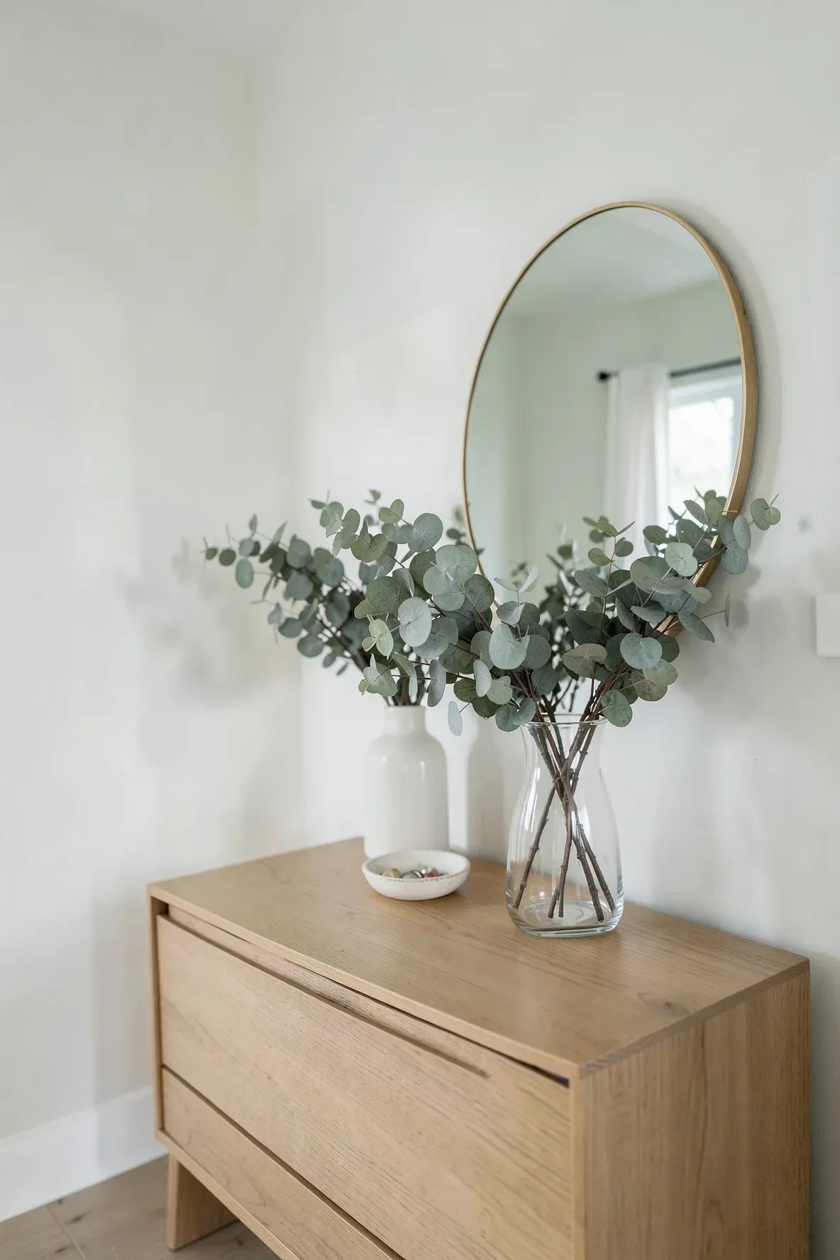 A clear glass vase with dried eucalyptus on a wooden dresser in a minimalist green bedroom.