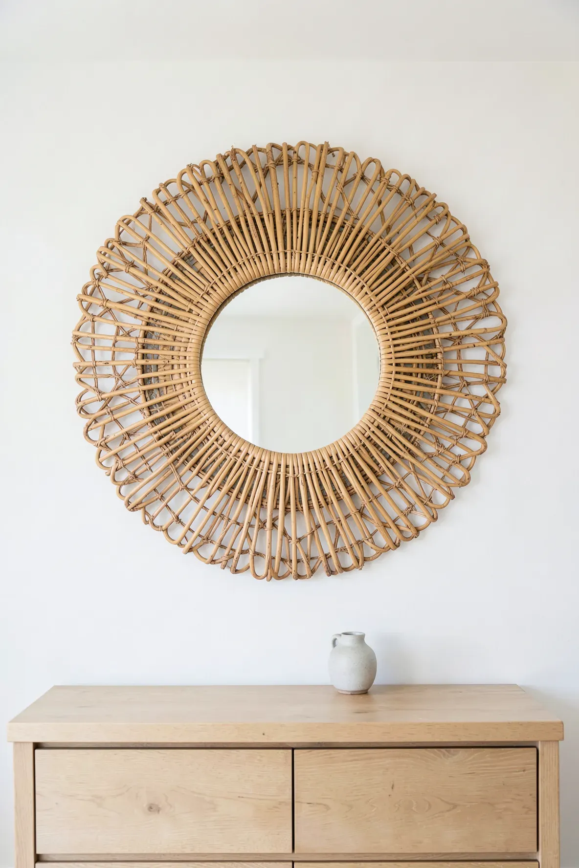 A decorative rattan sunburst mirror hanging on a white bedroom wall above a wooden dresser.