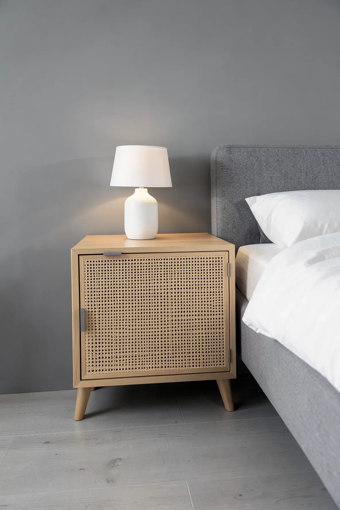 A nightstand with cane webbing and matte grey handles next to a grey bed.