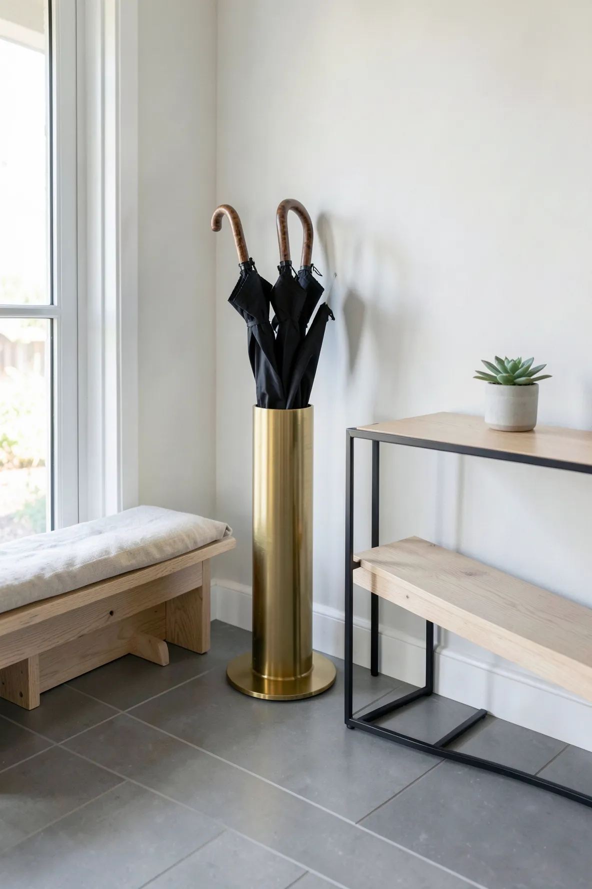 A modern brass umbrella stand tucked neatly next to a wooden entryway bench.