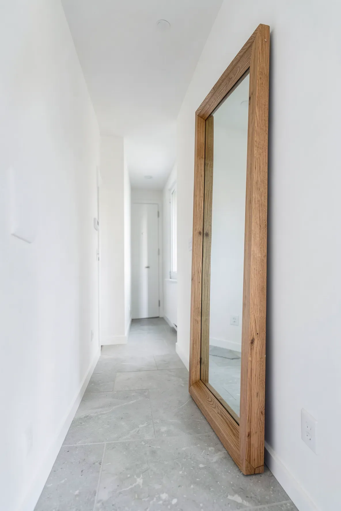A large oak wood frame mirror leaning against a white wall in a narrow, bright modern hallway.
