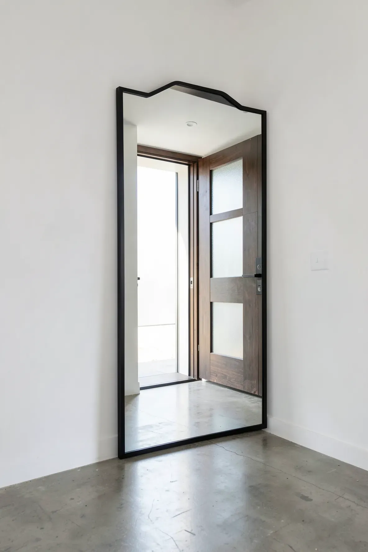 A minimalist black metal mirror reflecting bright sunlight from an open front door in a modern home.