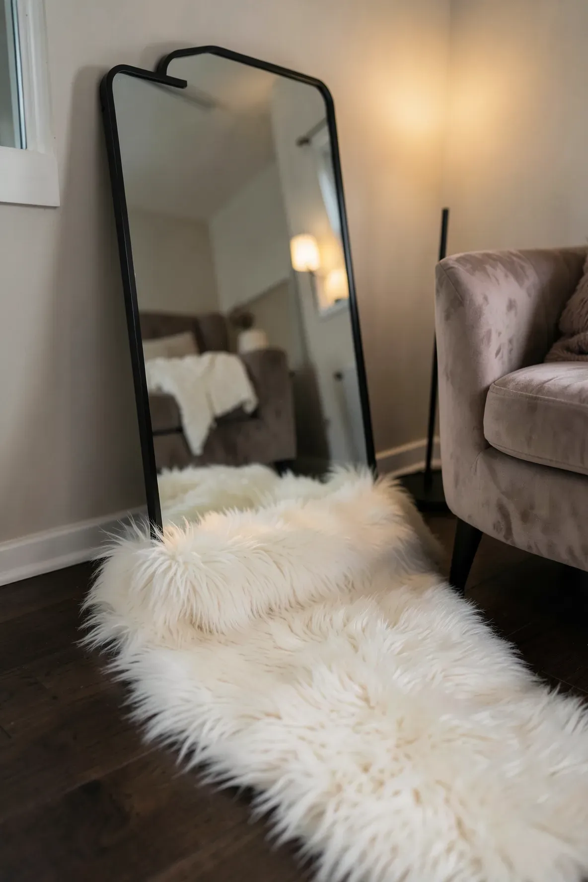 A black standing mirror with a fluffy white sheepskin rug at its base in a cozy room.