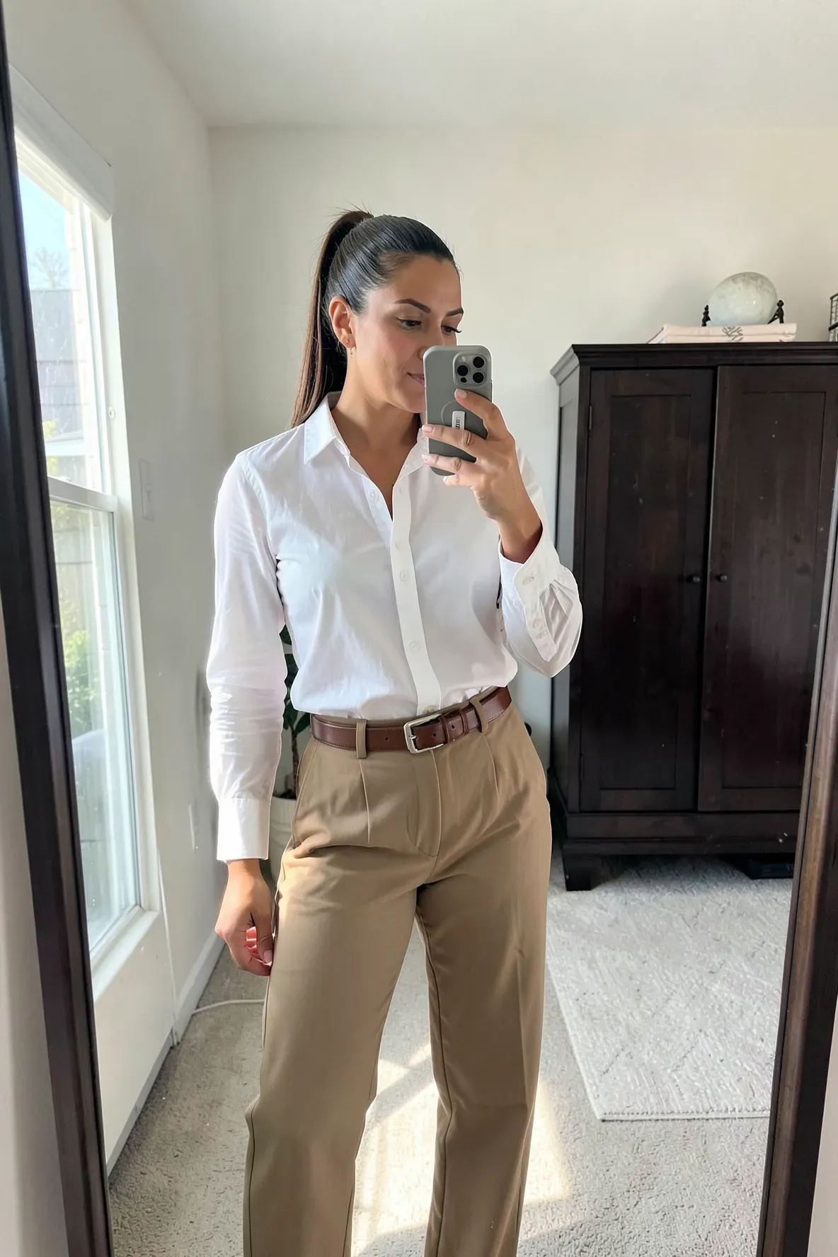 A woman in a white shirt and tan trousers taking a mirror selfie in a bright bedroom.