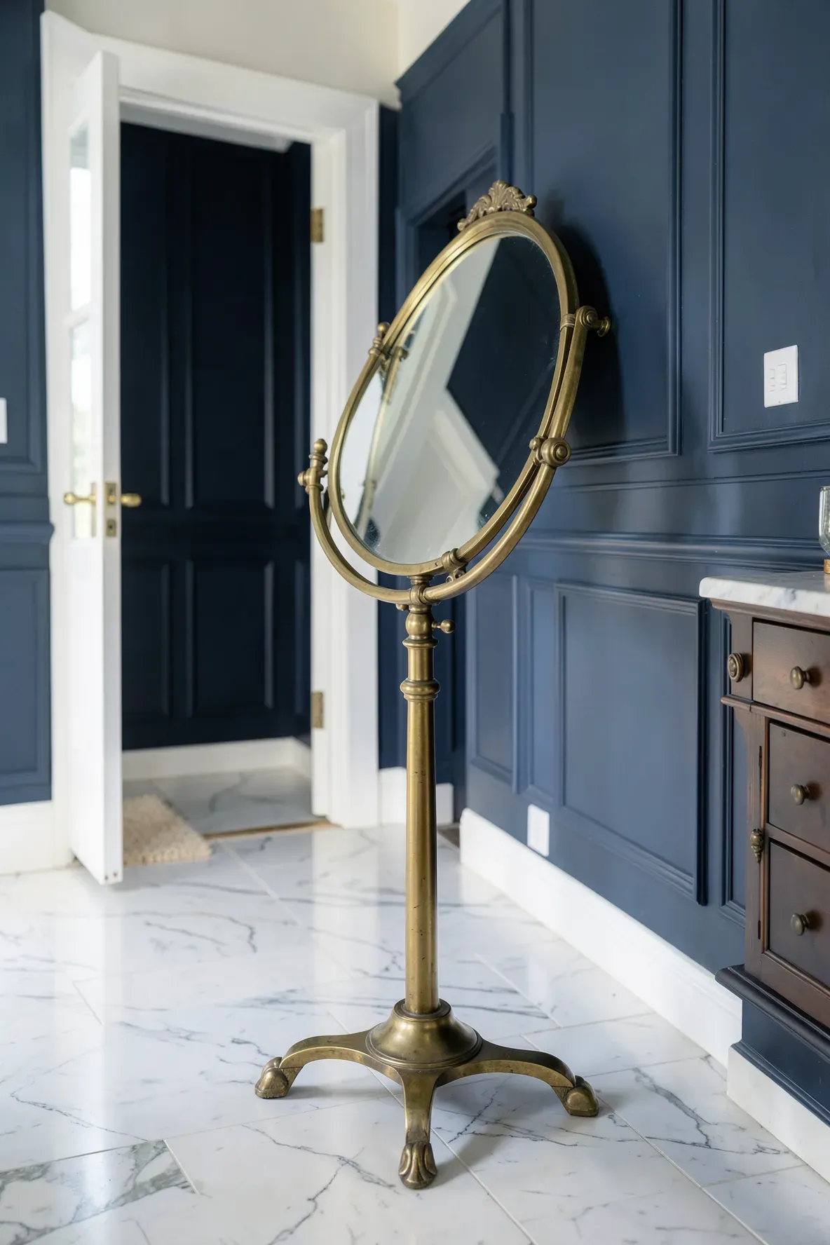 An adjustable antique brass pivot mirror standing on a marble floor in a navy blue foyer.