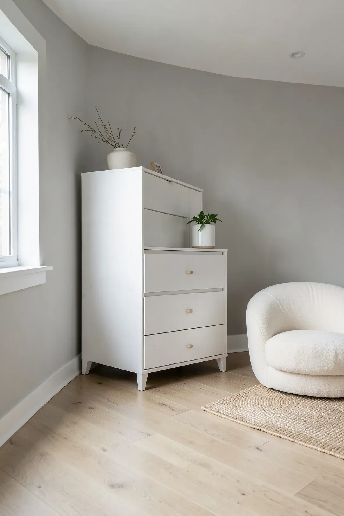 A tall white vertical dresser in a sunlit modern nursery with oak floors and a cream glider.