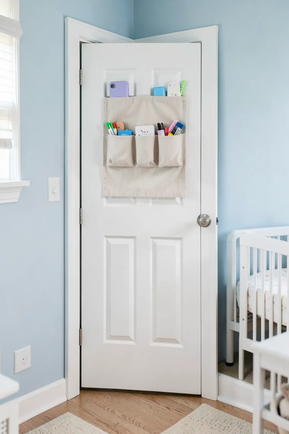 A white canvas over-the-door organizer hanging on a nursery door, holding baby socks and hats.