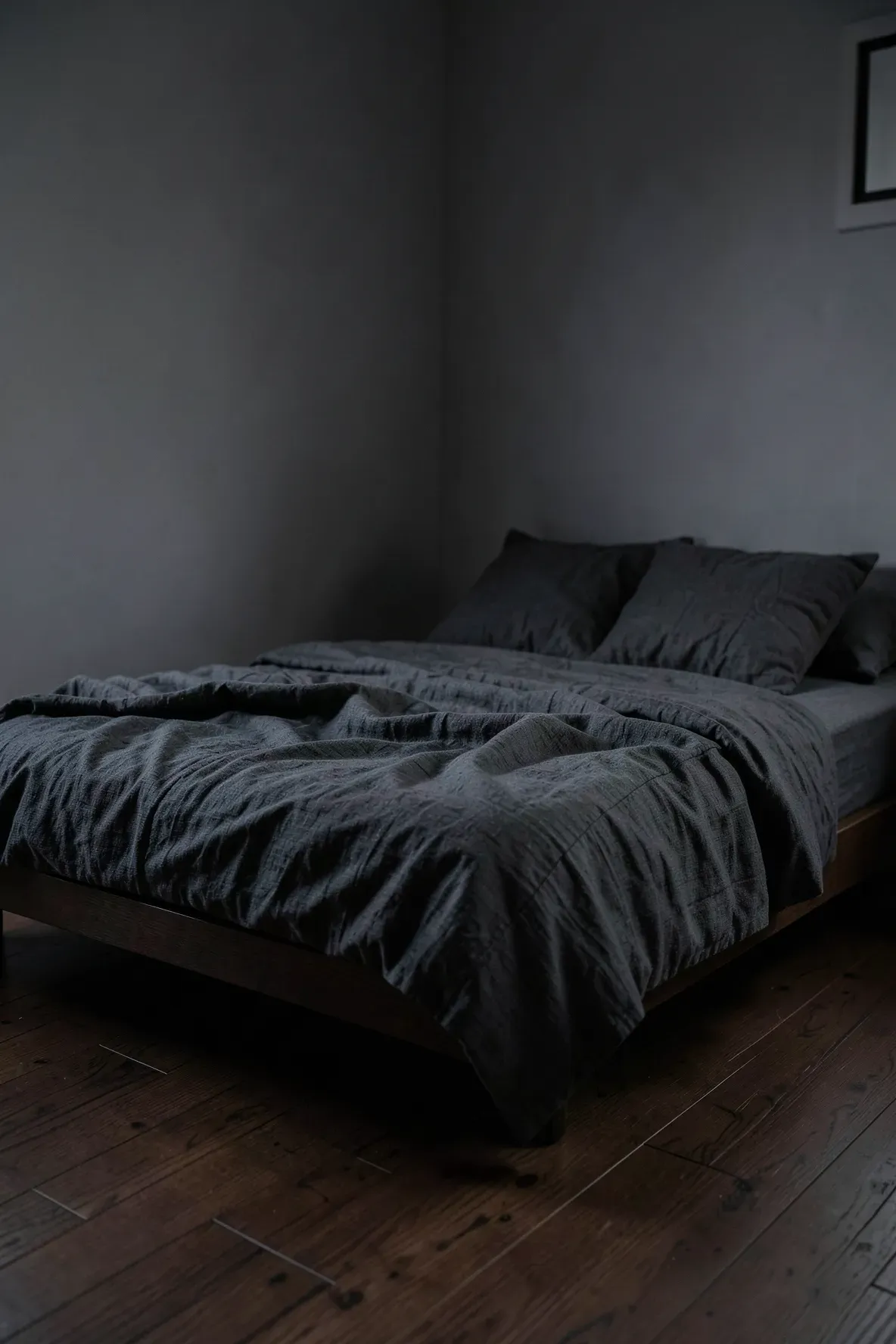 A low-profile bed with rumpled charcoal linen bedding in a dark, moody minimalist bedroom.