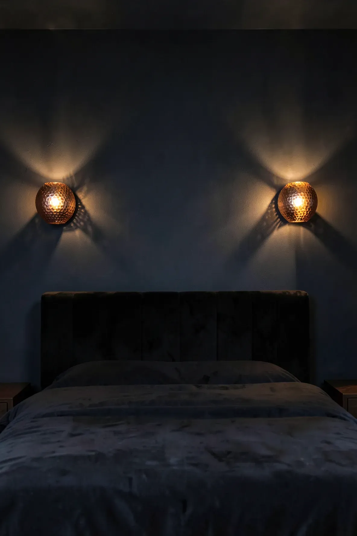 Hammered copper sconces glowing warmly against a dark wall in a moody, elegant bedroom.