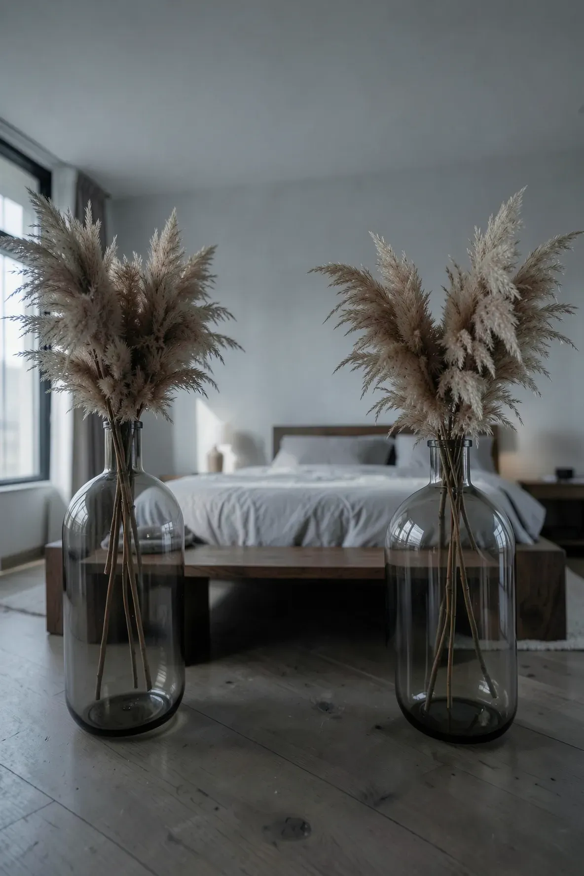 Dried pampas grass in smoked glass vases within a moody, minimalist bedroom setting.