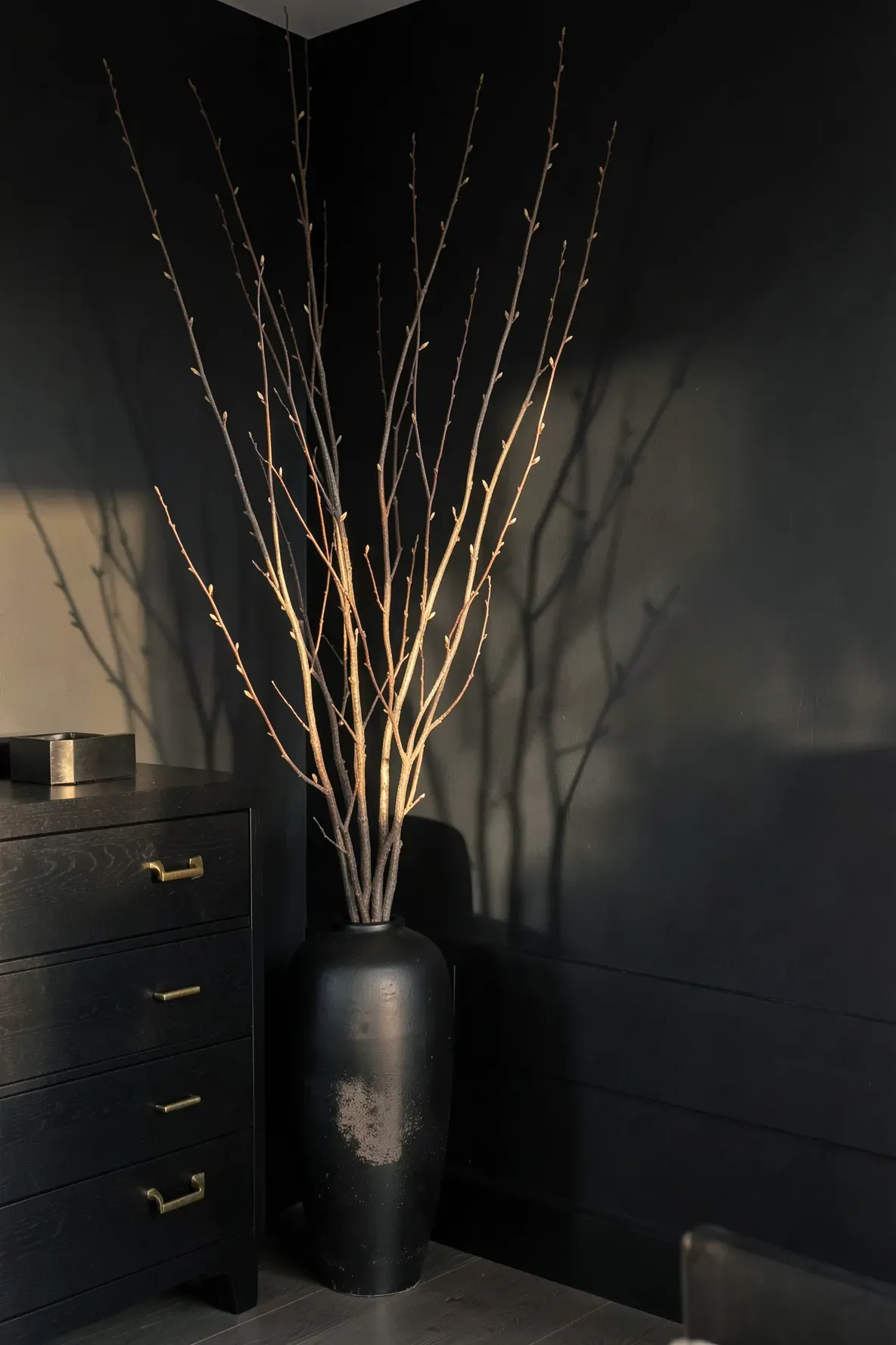 A tall matte black vase with bare willow branches in the corner of a dark, minimalist bedroom.