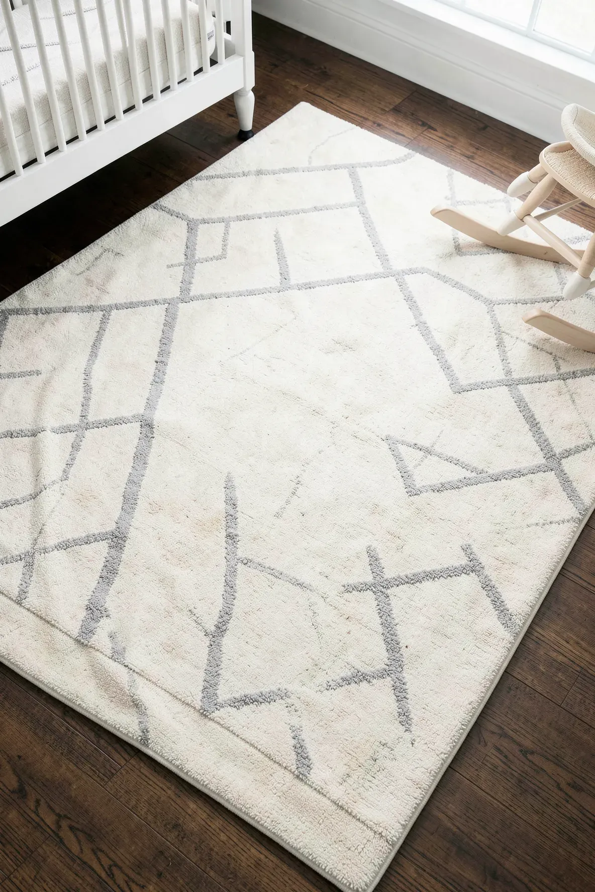 A cream and grey geometric washable rug on a dark wood nursery floor.