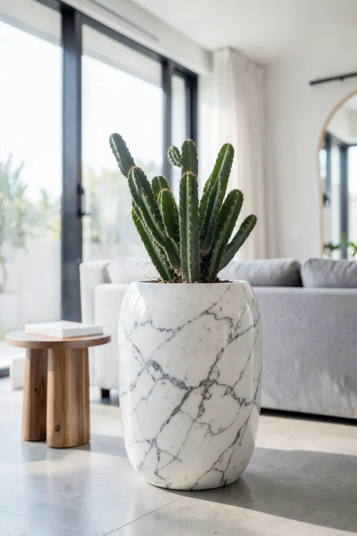 A tall green cactus in a heavy white marble vase on a wooden table in a bright modern room.