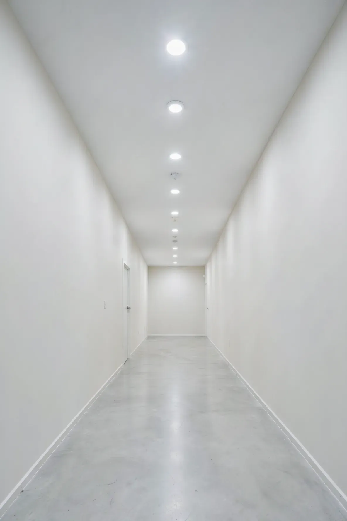 Evenly spaced recessed LED lights on the ceiling of a long narrow hallway providing bright light.
