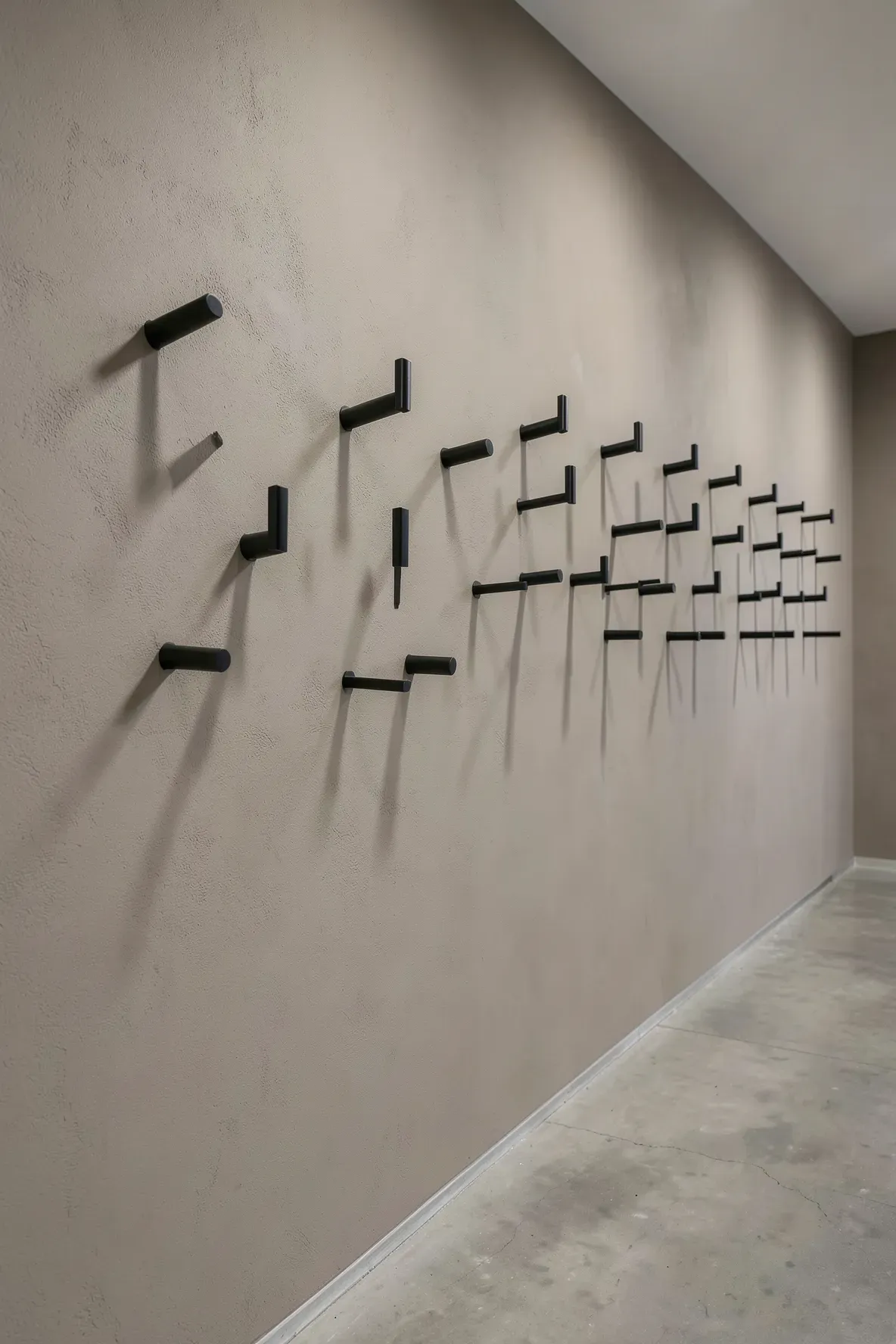 Matte black metal hooks arranged in a staggered artistic pattern on a taupe hallway wall.