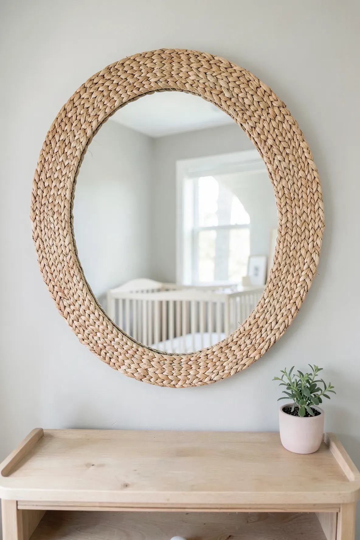 A round mirror with a woven seagrass frame hanging on a light grey wall in a bright baby room.