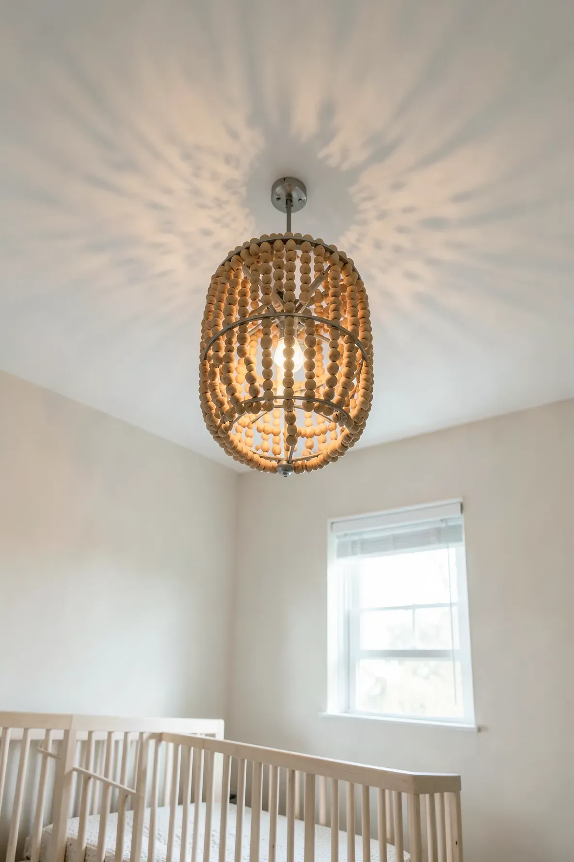 A decorative beaded wooden chandelier hangs in a calm, neutral-toned baby bedroom.