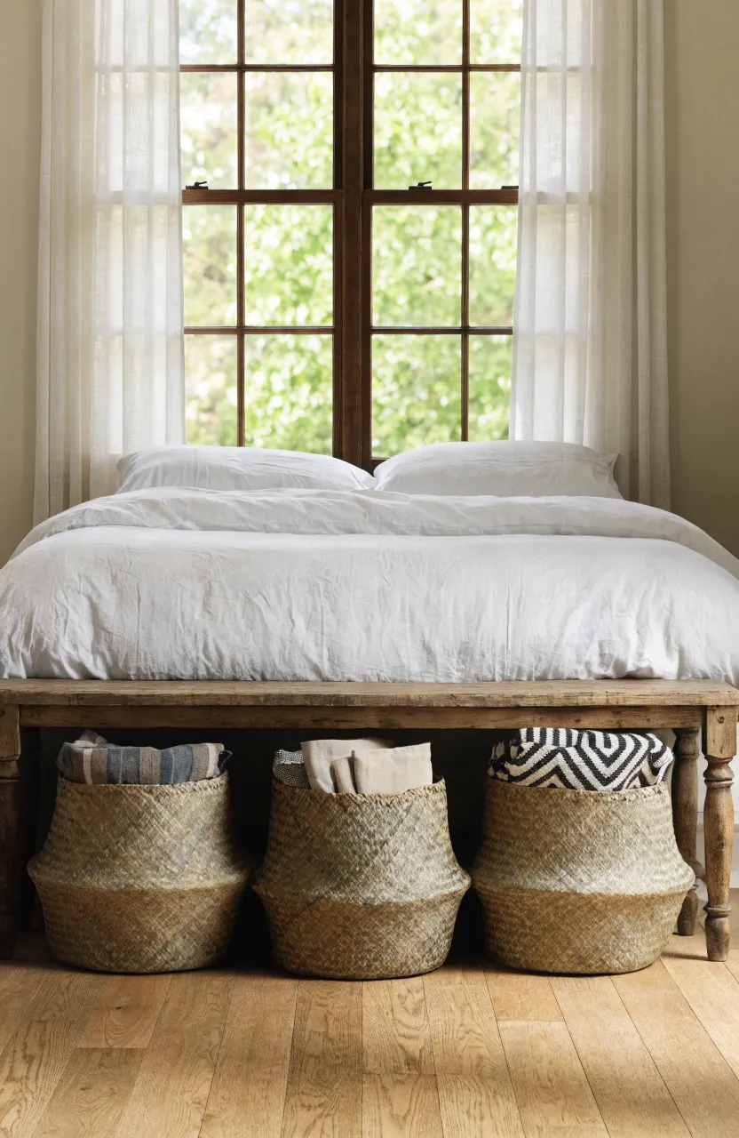 Woven seagrass baskets tucked under a wooden bench at the foot of a bed in a neutral earthy bedroom.