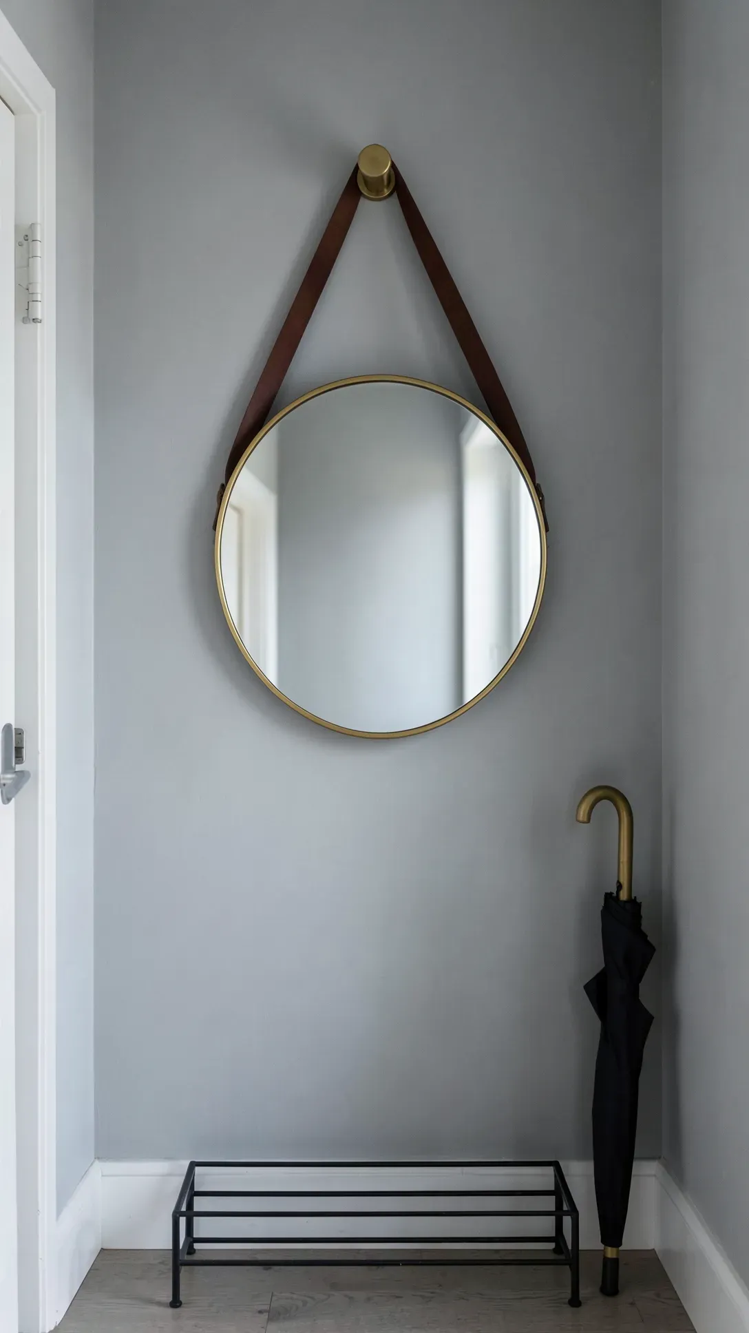 A round mirror hanging from a leather strap and brass peg in a modern, compact entryway.