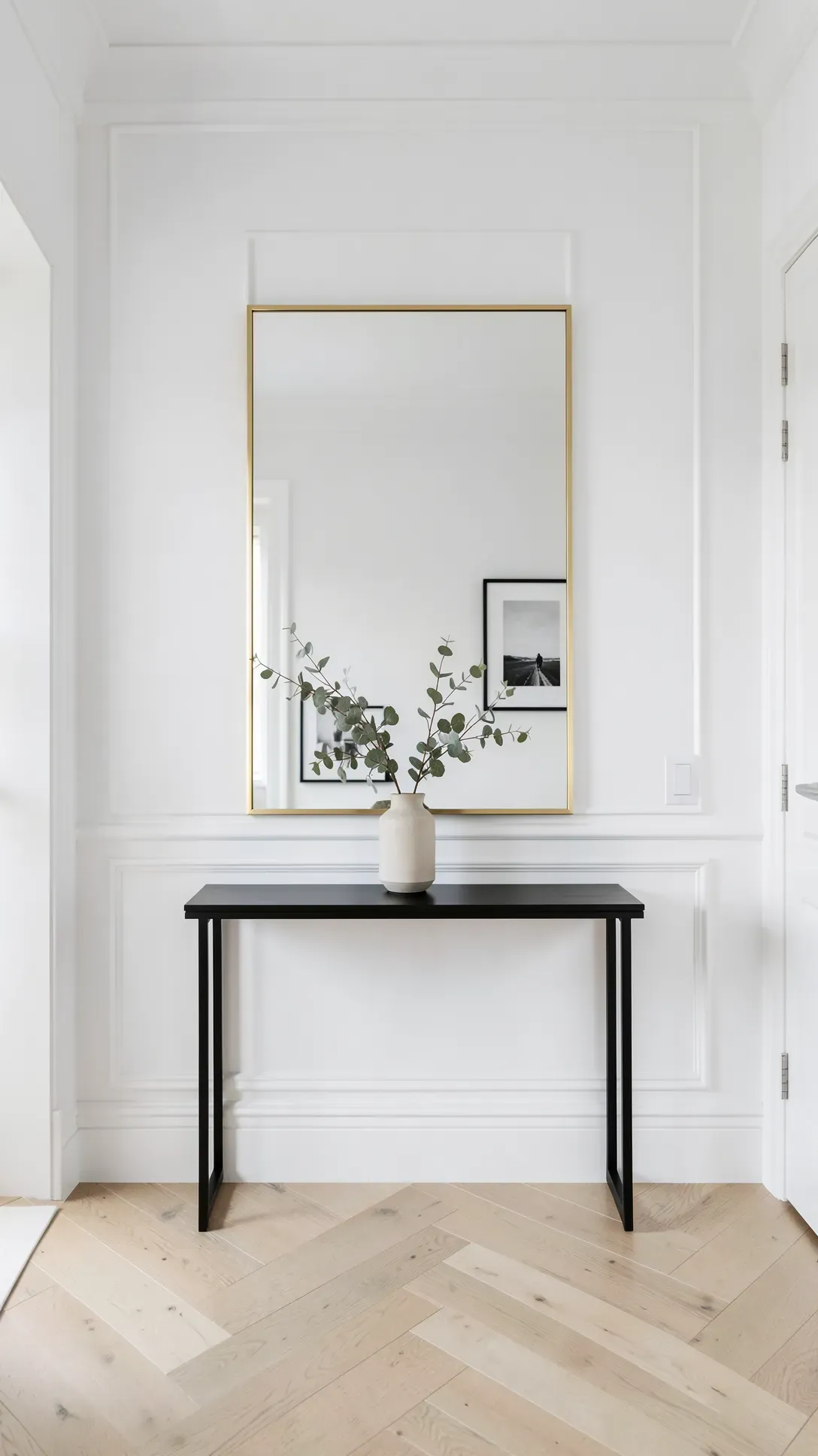 A minimalist entryway featuring a thin brass-framed mirror above a black console table in a bright foyer.